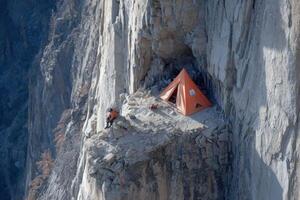 Individual sits near bright orange tent perched on rocky ledge, surrounded by towering cliffs and expansive valley views under clear skies photo