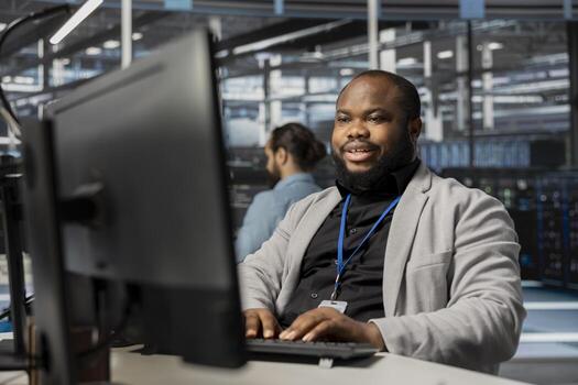 Cheerful data center employee working to update rackmounts software, configuring network virtualization. Upbeat african american man in server room verifying mainframes settings photo