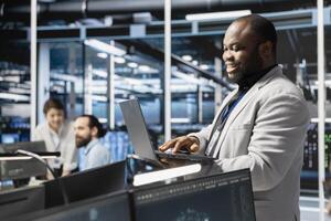 Happy data center facility admin using laptop to update supercomputers software, configuring network virtualization. Jolly server hub IT professional using notebook to verify equipment settings photo