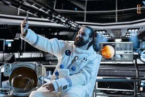 Astronaut wearing spacesuit inside futuristic spacecraft doing selfie with smartphone. Smiling spaceman wearing equipment doing photos with mobile phone during space travel operations