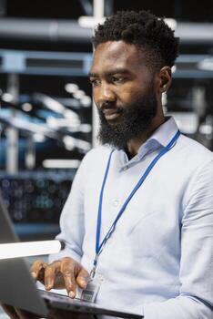 Computer scientist in server room uses laptop to implement and manage data backup solutions. African american man in data center identifying and resolving technical errors, ensuring efficient recovery photo