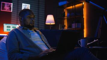 Skilled african american man multitasking on laptop in cool living room, connected to the internet while managing freelance work and remote responsibilities. Browsing web on bean bag. Camera A. photo
