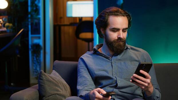 Smiling person on couch picking movie on streaming service using remote and texting on mobile phone. Happy man watching shows on television, zapping though channels, chatting on smartphone, camera A photo