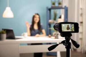 Behind the scenes of beauty vlogger filming tutorials, with a camera on a tripod focusing on her seated at a desk against a blue backdrop. Female content creator waving hello towards recording device. photo