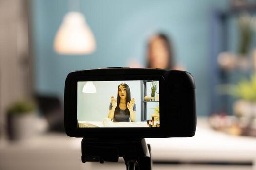 A digital camera capturing a beauty vlogger in action. Asian woman blogger is seen through the device screen, using expressive hand gestures while discussing makeup products in a studio setup. photo