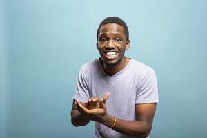 African american man with proud expression leaning forward and clapping his hands, happy with the good news. Young black guy standing against blue background, looking at camera and giving an applause. photo