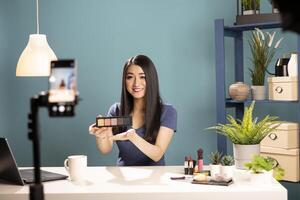 Friendly asian vlogger presenting an eyeshadow palette towards her cellphone camera, recording a . She demonstrates makeup techniques, holding a product while filming for an online tutorial. photo