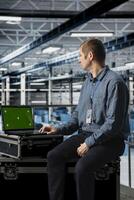 Engineer using chroma key laptop in server hub, conducting hardware installations and upgrades. Data center employee using green screen notebook, implementing server virtualization solutions photo