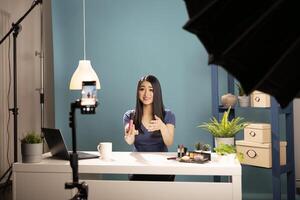 Beauty content creator records with a cellphone in studio, engaging her audience with a cosmetic review. The scene includes blue background, lighting equipment, and mobile device on a tripod. photo