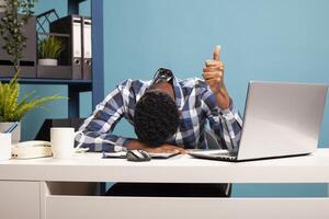 Young black businessman resting head on desk and giving thumbs up, exhausted but celebrating success. Sleepy office worker showing approval sign and napping on table after tiring workday. photo
