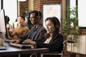 Smiling female employee wearing wired headphones works attentively at the office desk. Caucasian woman in wheelchair with a headset communicates with online clients while entering data on her computer. photo