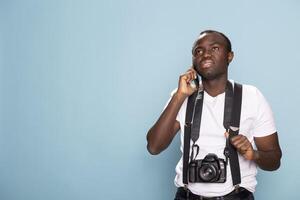 Young black man with a DSLR camera around his neck talking on his mobile device. Male photographer wearing suspenders and standing confidently in studio as he speaks with a client on his cellphone. photo