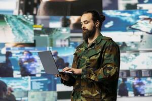 Military officer reviewing data from surveillance and works on tactical operations, tech driven mission control hub. Soldier uses threat detection electronic systems for national interests. photo