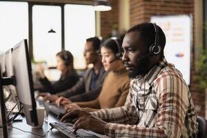 Portrait of african american customer support agent at his computer with coworkers nearby. Focused call center operator typing on his keyboard while listening to a client through his wired headphones. photo