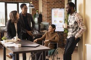 Multiethnic colleagues gather around a desk, taking a break and sharing stories in a relaxed office setting. Diverse group of employees stands, chatting with their asian coworker in a wheelchair. photo