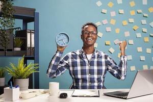 Happy businessman with a clock in hand, celebrating for finishing project presentation before deadline. Cheerful male manager raising fist with excitement, feeling great about end of workday. photo