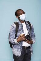 Cautious male photographer standing confidently with his camera and coffee, wearing face mask in blue background studio. He displays readiness for a photoshoot while staying protected against covid 19. photo