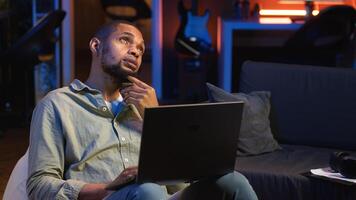 African american man working remotely from his modern loft on laptop, focusing on tasks completion while he lays on a bean bag. Blending productivity and relaxation, telework. Camera B. photo