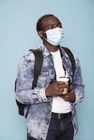 African american man in denim jacket and face mask holds his dslr camera and coffee cup, ready to meet his photography clients. Young photographer gazes upward while standing against isolated backdrop. photo