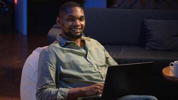 Focused black person multitasking on laptop in stylish loft space, connected to the internet while managing daily freelance tasks and remote responsibilities. Browsing web on sofa. Camera B. photo