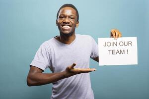 Enthusiastic recruiter looking at camera, holding a paper, inviting candidates to join his team. Smiling african american man pointing at hiring sign, promoting job openings in an isolated background. photo