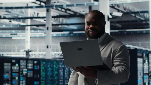 Server room worker auditing systems for vulnerabilities using software on laptop. Engineer walking in data center doing backups and recovery protocols using notebook, camera B photo