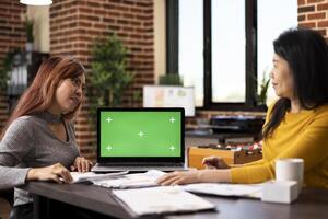Asian female colleagues reviewing startup company documents at office desk as a digital device displays blank mockup template. Laptop with greenscreen is placed on table between women in a discussion. photo