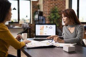 Asian woman advisor with pen in hand, consulting her coworker using laptop. Female professionals working together at office desk, utilizing digital device and documents in their business discussion. photo