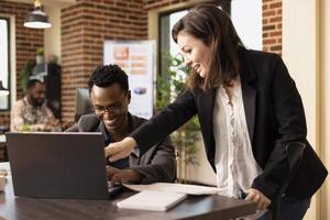 Black man sharing a laughter with his caucasian female coworker at the office desk while having a business discussion. Cheerful multiethnic colleagues having a meeting, reviewing reports on laptop. photo