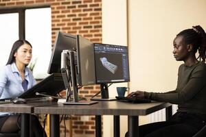 African american developer designing 3d machinery equipment using technical CAD software on computer. Multiethnic female engineers seated in creative agency office, working on various projects. photo