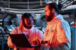 Teamworking spaceship engineers using laptop to fix security breach causing overload warning. Space station colleagues using notebook, repairing critical system error triggering emergency lights photo