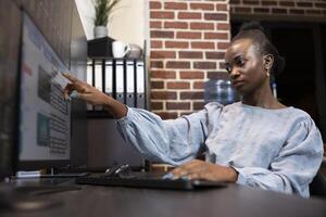 Black female finance analyst monitoring forex data on multiple screens, pointing at her computer, assessing stock movements in a professional brick walled workspace. Focused woman working at her desk. photo