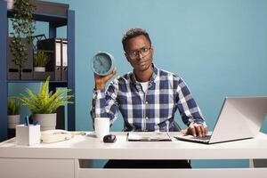 Project team leader sitting at desk with a clock in his hand, showing annoyance at his employees coming late to work. Black male manager using laptop, displeased by late comers, giving time evidence. photo