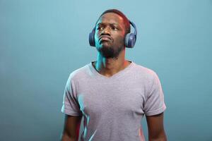 Black man focused on listening to podcast through his wireless headset in a blue background studio. African American male model standing and looking up with serious expression, using his headphones. photo