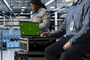 Close up of data center employee using green screen notebook, managing automation scripts for optimized operations. Engineer using mockup laptop in server farm, maintaining optimal system performance photo
