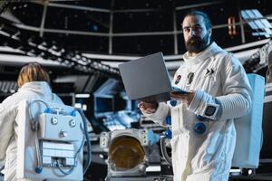 Astronaut reviewing spacecraft diagnostics on laptop to ensure safe operation during long distance space travel. Spaceman uses notebook to monitor system stability during cosmic journey photo