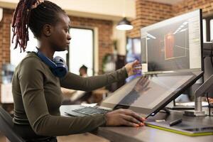 Determined black woman in a creative agency developing innovative digital art using internet based modeling tools. Young female editor seated at desk, focusing on a 3D design on her device screens. photo