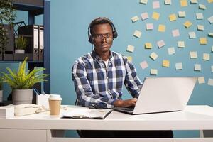 Male helpline operator seated at desk, looking at camera, struggling with fatigue from long work hours. Portrait of black man with crooked glasses and headset, appearing drained from overtime workload photo