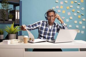 Joyful manager seated at desk, dancing and reviewing company information with paperwork and his laptop. Male analyst with headphones, flipping documents on clipboard and enjoying music on headphones. photo