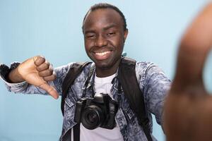 POV of male photographer standing and playfully giving a thumbs down against isolated background. African american vlogger with dslr camera and backpack recording himself as he gestures disagreement. photo