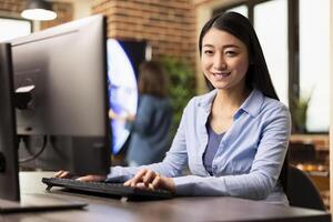 Young female employee sitting at desk in modern office, happily working on computer and looking at camera. Portrait of asian businesswoman typing on keyboard, engaged in work at creative workspace. photo
