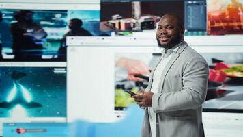 Portrait of smiling man browsing social networks feed on smartphone in front of videowall. Jolly african american person uses phone, floating icons highlighting interactive engagement online, camera A photo