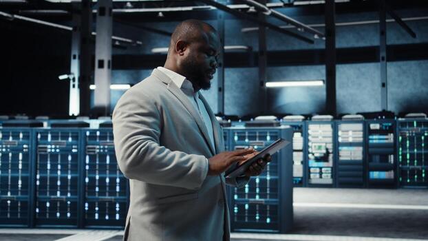 Manager walking in server hub using tablet to find firewall misconfigurations affecting rackmounts performance. Ad,om checking systems bottlenecks leading to sluggish data transfer rates, camera A photo