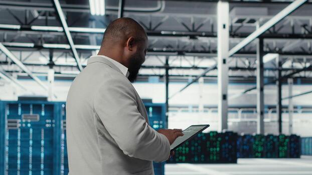 Cheerful server room engineer troubleshooting backend operations using software on tablet. Jolly data center programmer compiling software patches to fix bugs using device, camera B photo