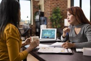 Female professionals sitting with documents and using data charts on tablet for comparisons in modern workspace. Businesswomen, one with coffee in hand, are having a project discussion in office. photo