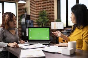Digital device displaying a blank chromakey template sits on the table alongside scattered paperwork, with two women having a business discussion. Strategy meeting between coworkers. photo