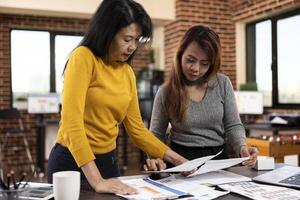 Focused asian consultants standing at desk, focused on company research documents in modern workspace. Businesswomen near table, looking at paperwork, reviewing and discussing in startup office. photo