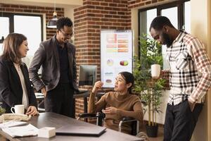 Multiethnic startup team standing and listening to their female coworker in wheelchair as she shares a hilarious story. Diverse group of employees bonding during their break in modern workspace. photo