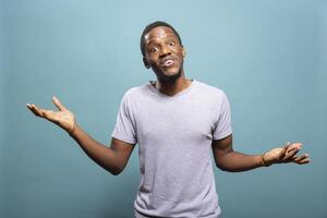 Young black man doing I dont know gesture in a studio, raising shoulders and expressing indecision. African american guy standing with open arms, shrugging in confusion against an isolated background. photo