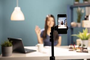 Closeup of a mobile device focused on a beauty influencer in the screen display. Asian woman waving at phone camera, ready to demonstrate makeup techniques to her vlogging channel. photo